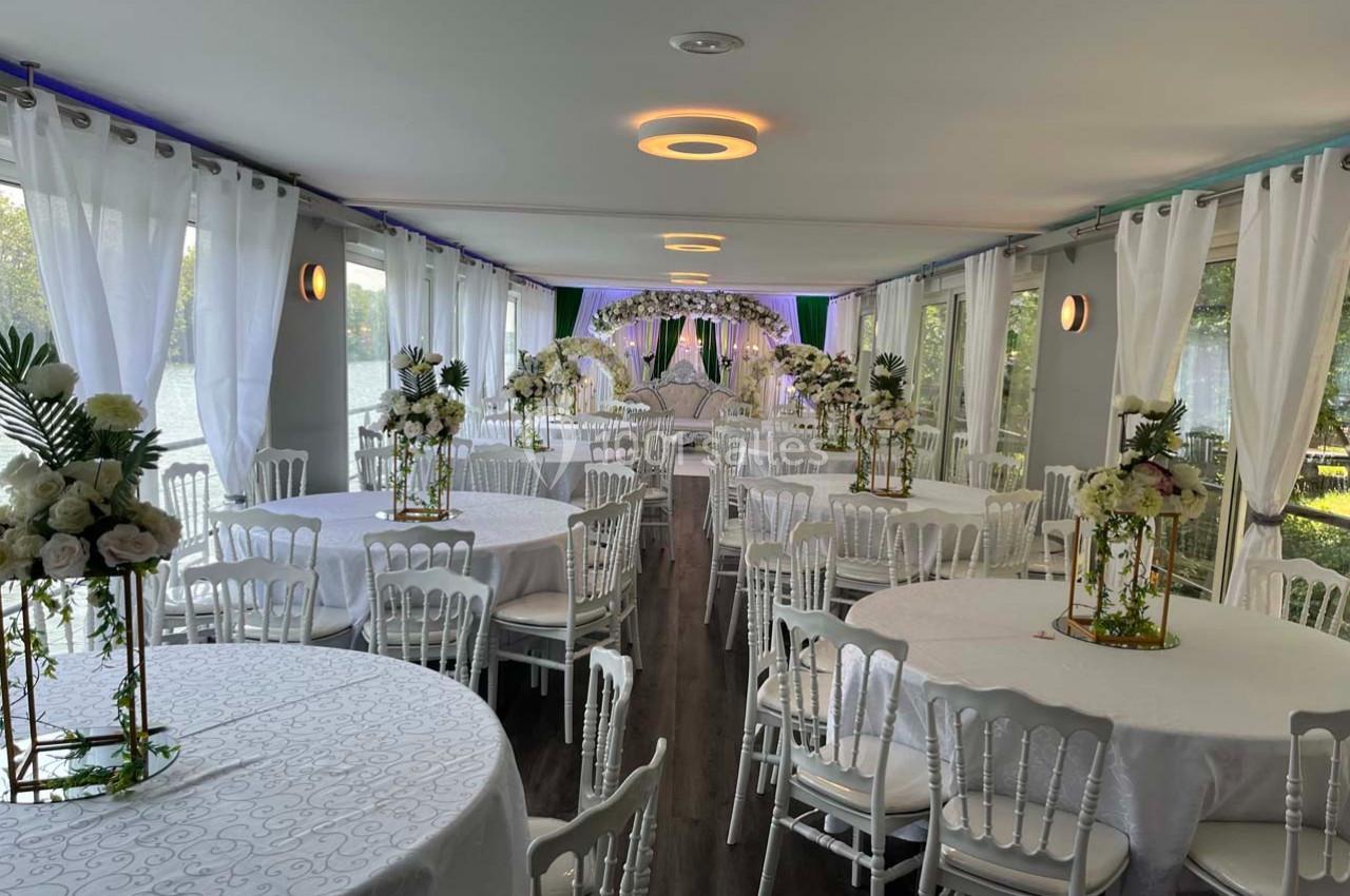 Salle de réception élégante avec tables rondes décorées de fleurs blanches et chaises blanches, éclairée par la lumière…