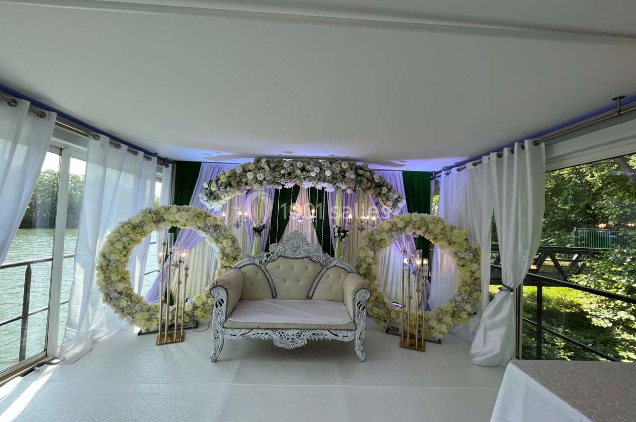 Canapé blanc décoratif entouré d'arcs floraux et de rideaux blancs dans un espace lumineux avec vue sur l'extérieur…