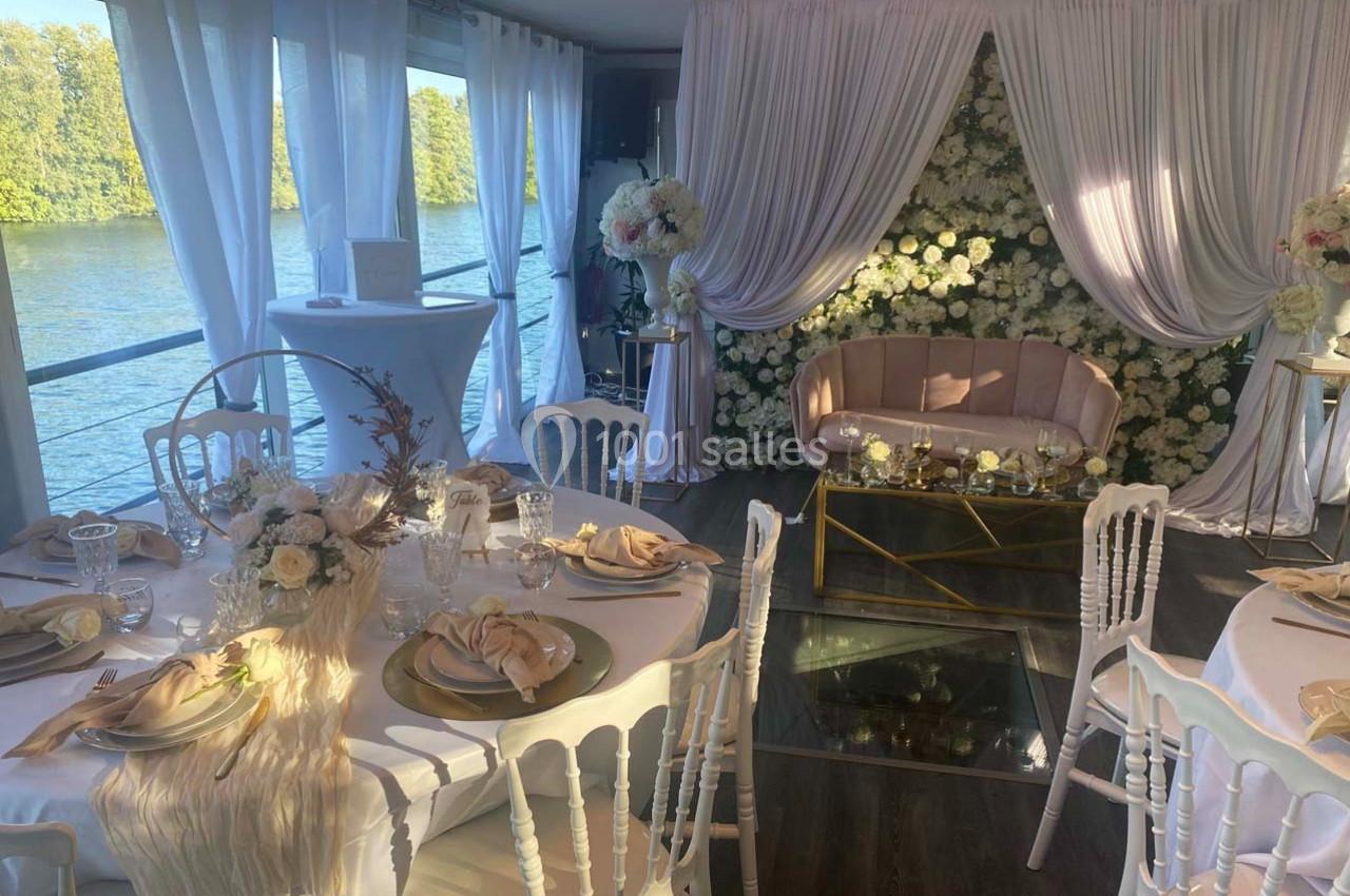 Salle décorée pour un événement avec des tables élégantes, des fleurs blanches et un canapé devant un mur floral.