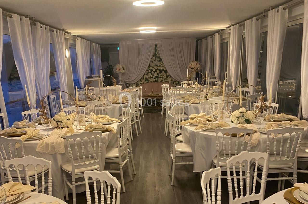 Salle de réception élégamment décorée avec des tables rondes, nappes blanches, chaises assorties et arrangements floraux.