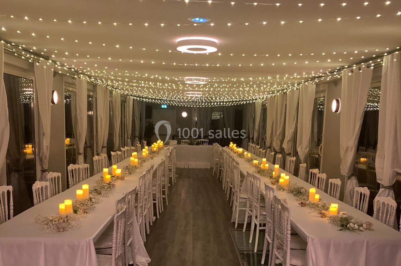 Salle de réception décorée avec des guirlandes lumineuses au plafond, tables alignées et bougies allumées.