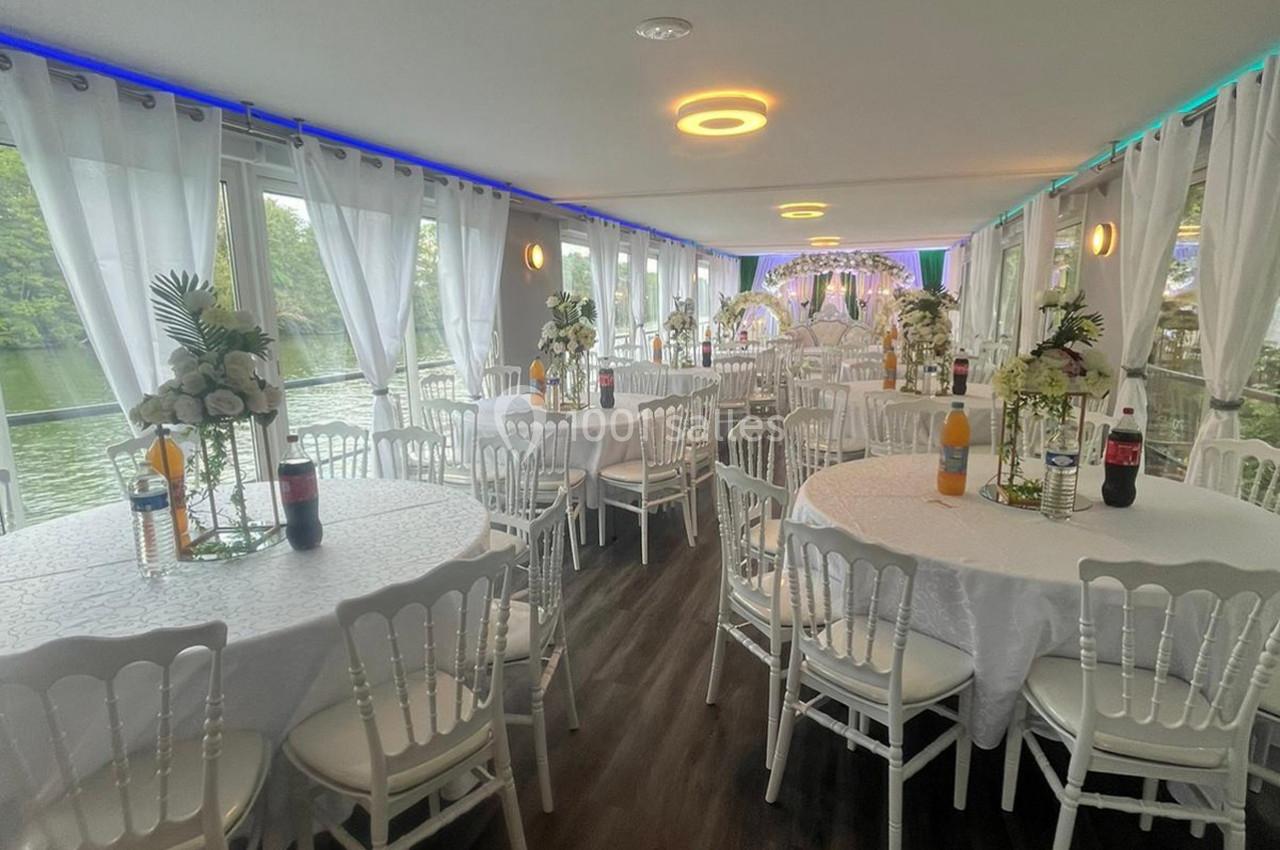 Salle de réception décorée avec des tables rondes, nappes blanches, chaises élégantes et vue sur l'extérieur verdoyant.
