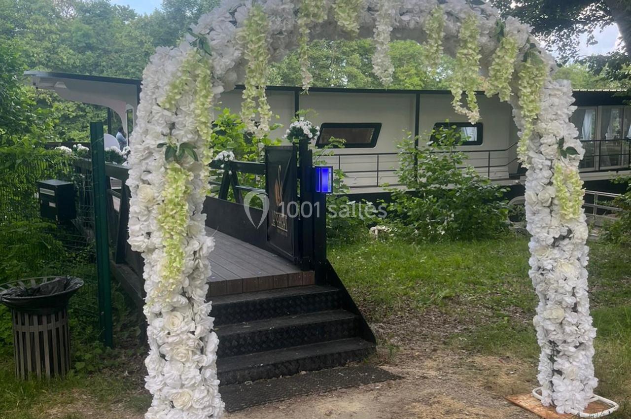 Arche décorée de fleurs blanches et vertes devant une passerelle menant à un bâtiment entouré de verdure.