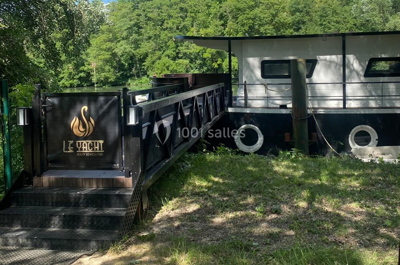 Passerelle en bois menant à une péniche noire amarrée, entourée de verdure et d'arbres.