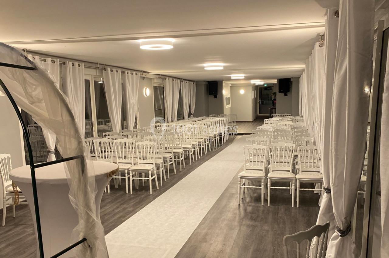 Salle de réception décorée avec des chaises blanches alignées, un chemin central et des rideaux blancs aux fenêtres.
