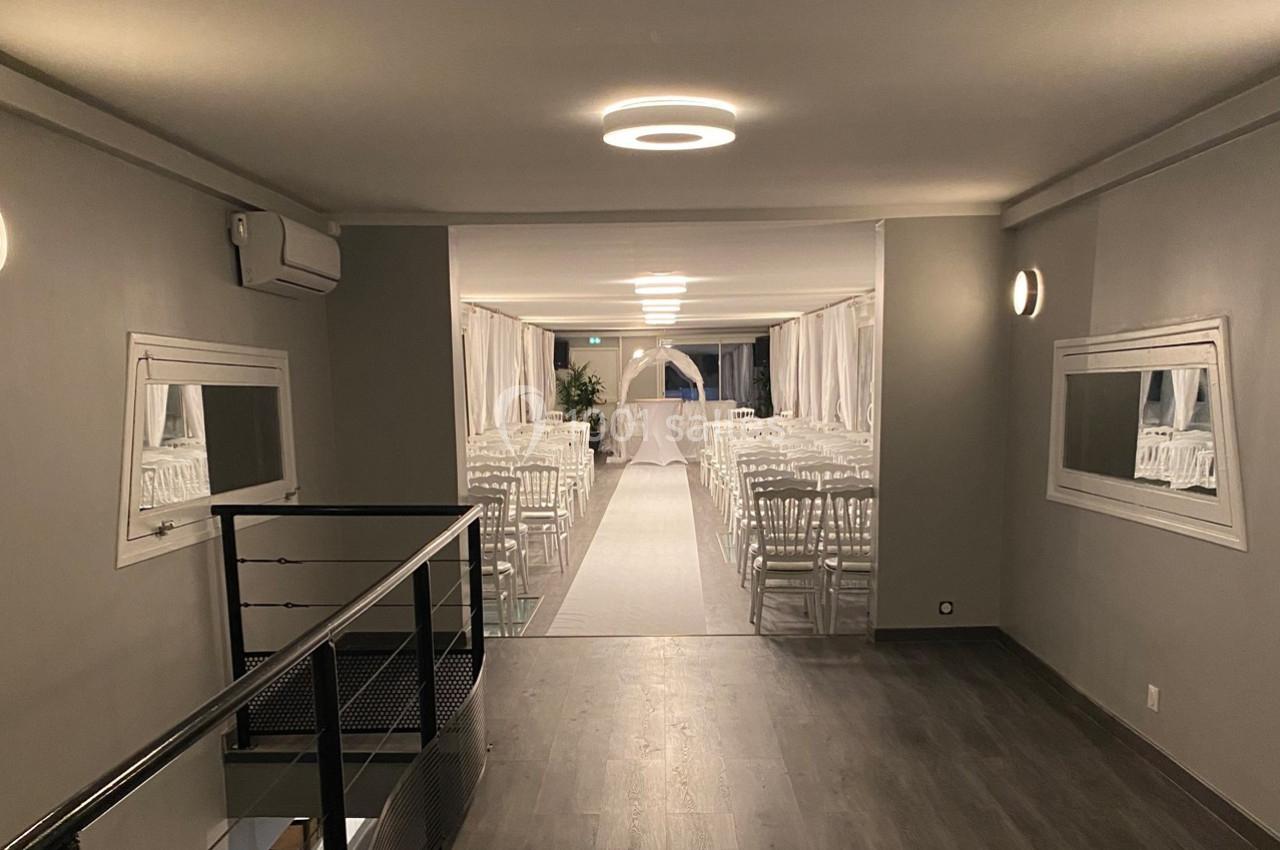 Salle lumineuse aménagée pour une cérémonie, avec des rangées de chaises blanches et une arche décorative au fond.