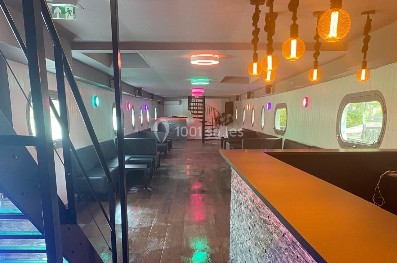 Intérieur d'un espace moderne avec bar, éclairage coloré, escaliers métalliques et hublots sur les murs.