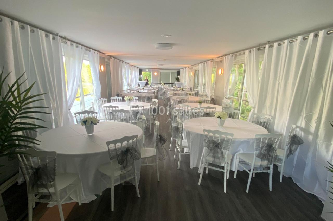 Salle de réception lumineuse avec tables rondes dressées de nappes blanches et chaises décorées de nœuds gris.