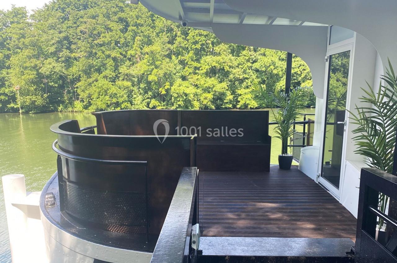 Terrasse en bois d'un bateau avec garde-corps courbé, entourée d'arbres et donnant sur une rivière calme.