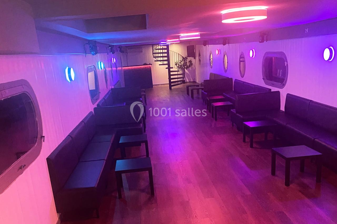 Salle intérieure avec éclairage coloré, canapés noirs alignés le long des murs et tables basses sur un sol en bois.