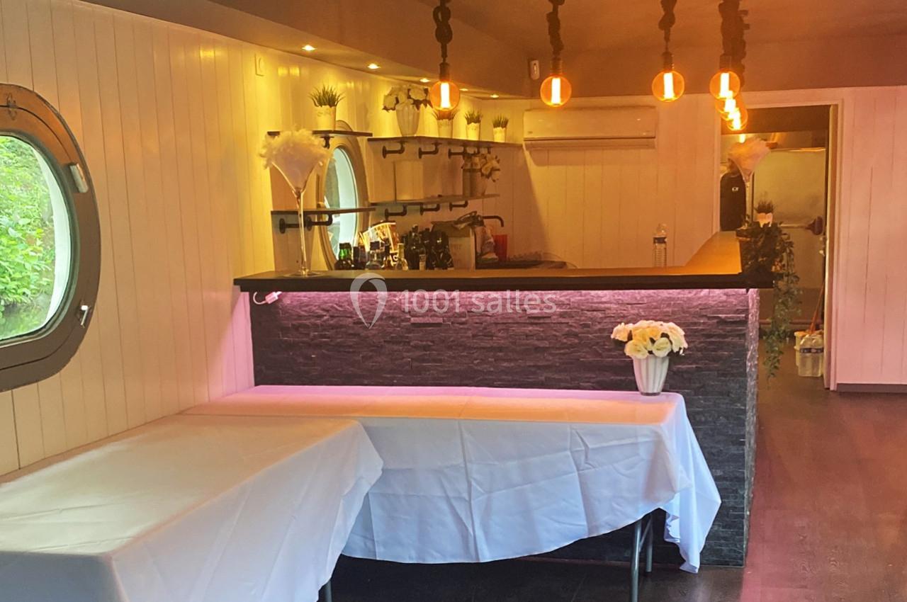 Intérieur d'un espace aménagé avec un bar en pierre, des tables couvertes de nappes blanches et des luminaires suspendus.