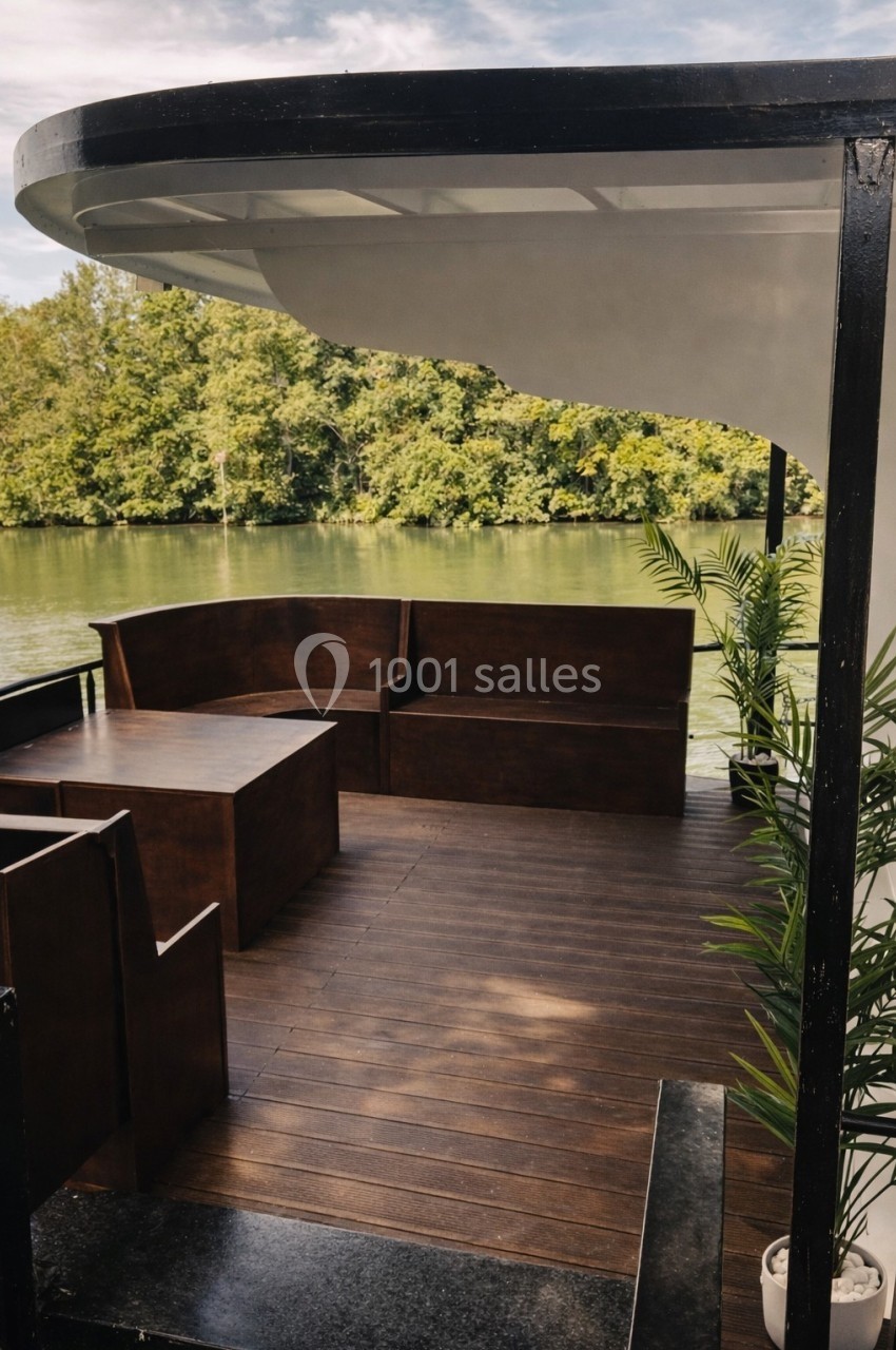 Terrasse en bois avec des bancs et des plantes, donnant sur une rivière bordée d'arbres sous un ciel partiellement nuageux.