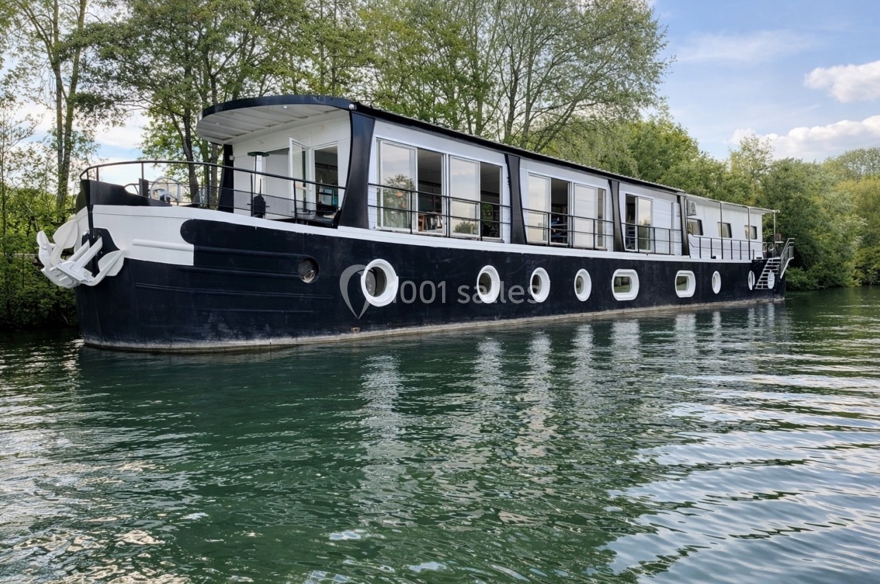 Péniche noire et blanche amarrée sur une rivière entourée d'arbres sous un ciel partiellement dégagé.
