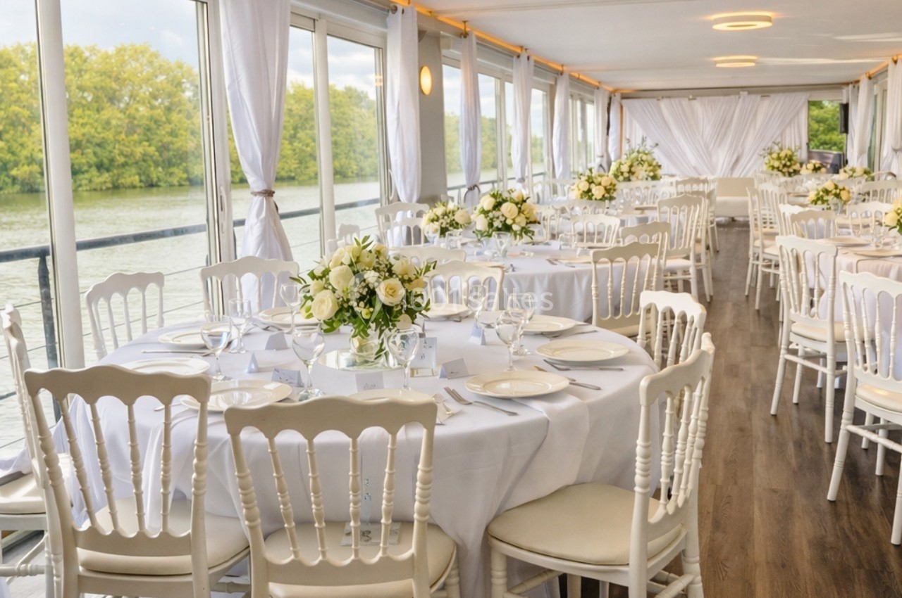Salle de réception lumineuse sur un bateau, décorée de tables rondes avec nappes blanches et bouquets de fleurs.