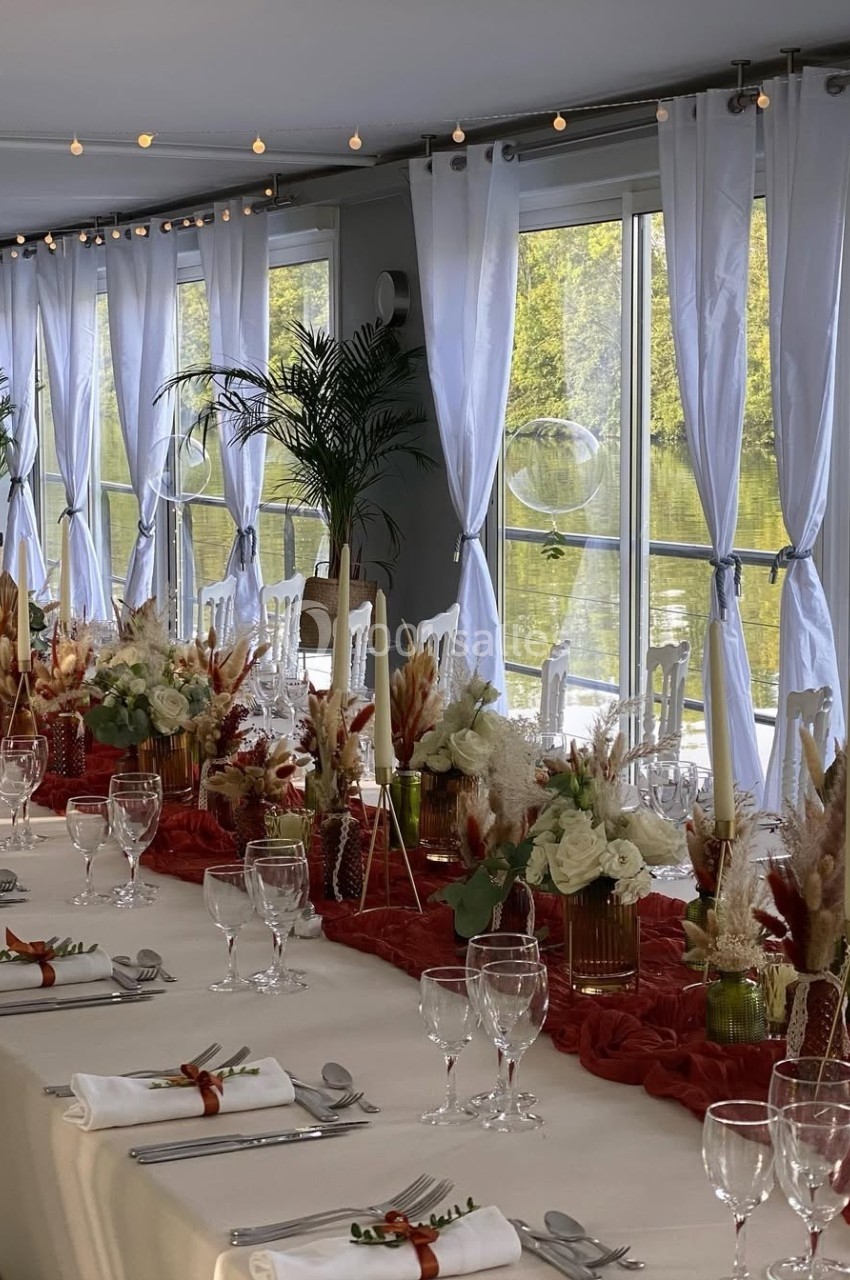 Table élégamment dressée avec nappes blanches, décorations florales rouges et blanches, et vue sur un paysage verdoyant.