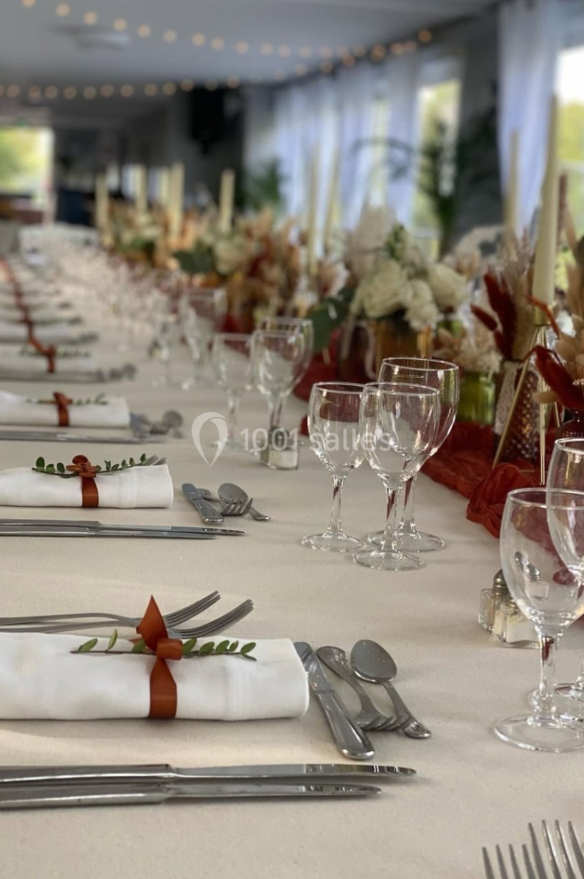 Table élégamment dressée avec nappes blanches, serviettes pliées, verres et décorations florales dans une salle lumineuse.