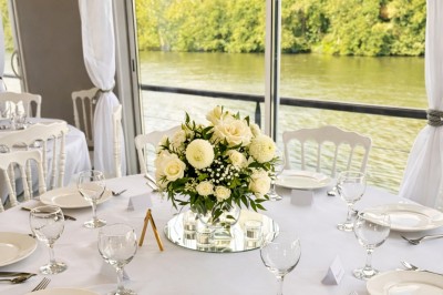 Table élégamment dressée avec nappes blanches, serviettes pliées, verres et décorations florales dans une salle lumineuse.