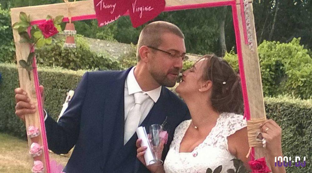 Un couple en tenue de mariage pose en souriant derrière un cadre décoré, dans un jardin verdoyant.