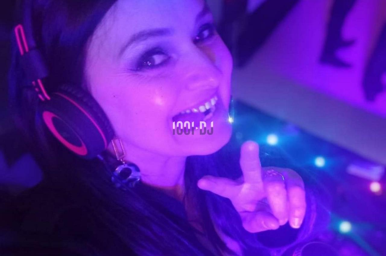 Femme portant un casque audio, souriante, faisant un signe de paix dans un environnement éclairé par des lumières colorées.