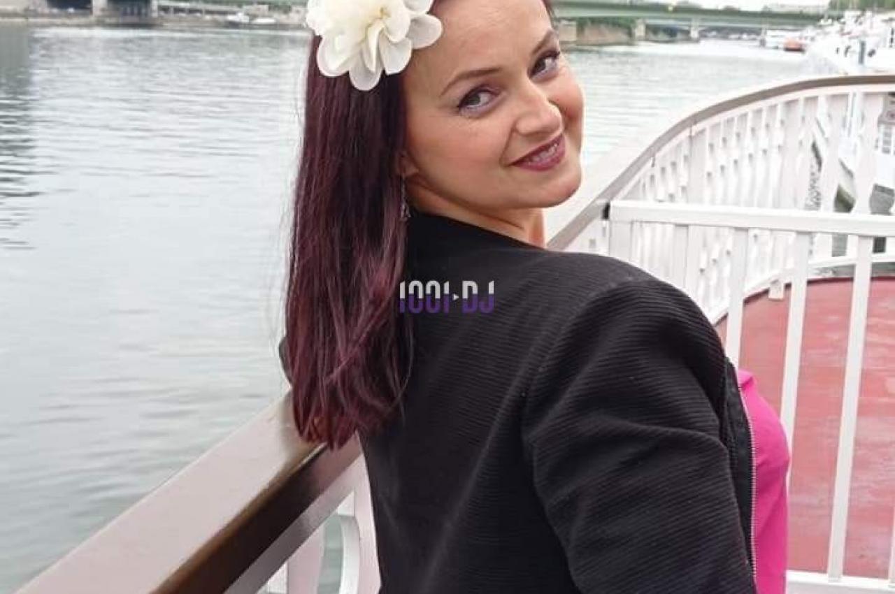Femme souriante avec une fleur blanche dans les cheveux, posant sur un pont avec la Tour Eiffel en arrière-plan.