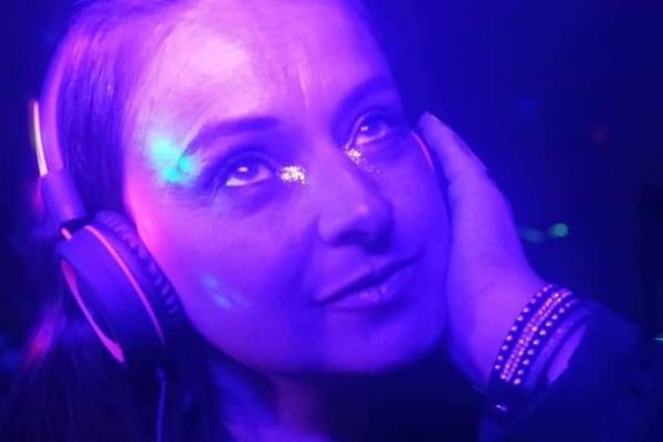 Femme portant un casque audio, éclairée par des lumières violettes, regardant vers le haut avec une expression songeuse.