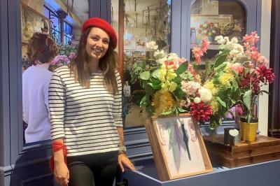 Femme souriante portant un béret rouge, debout près d'une vitrine décorée de fleurs colorées et de montres exposées.