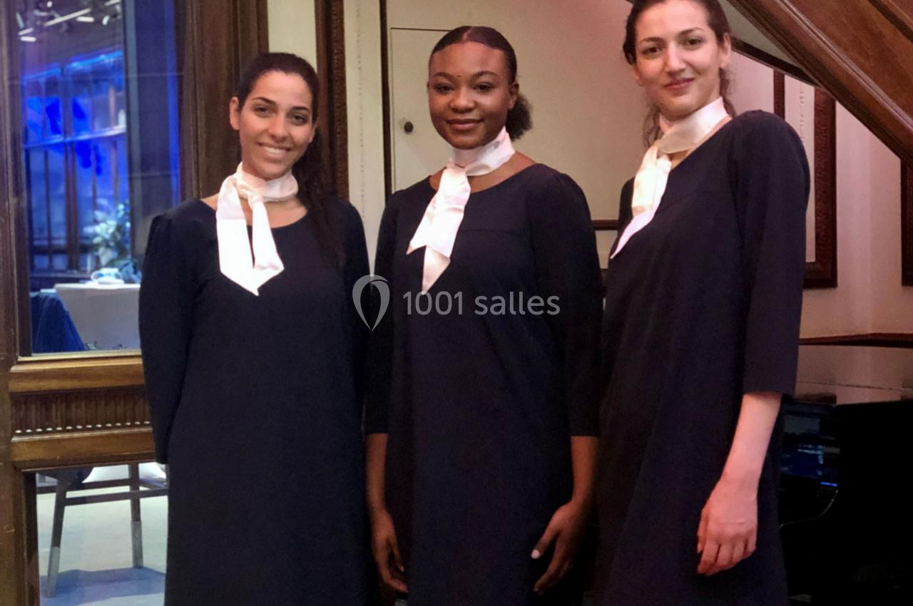 Trois femmes en tenue noire avec foulard blanc posent debout dans un intérieur élégant avec des cadres en bois.