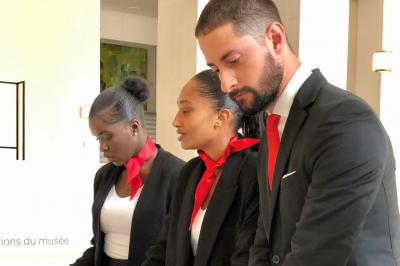 Femme souriante portant un béret rouge, debout près d'une vitrine décorée de fleurs colorées et de montres exposées.