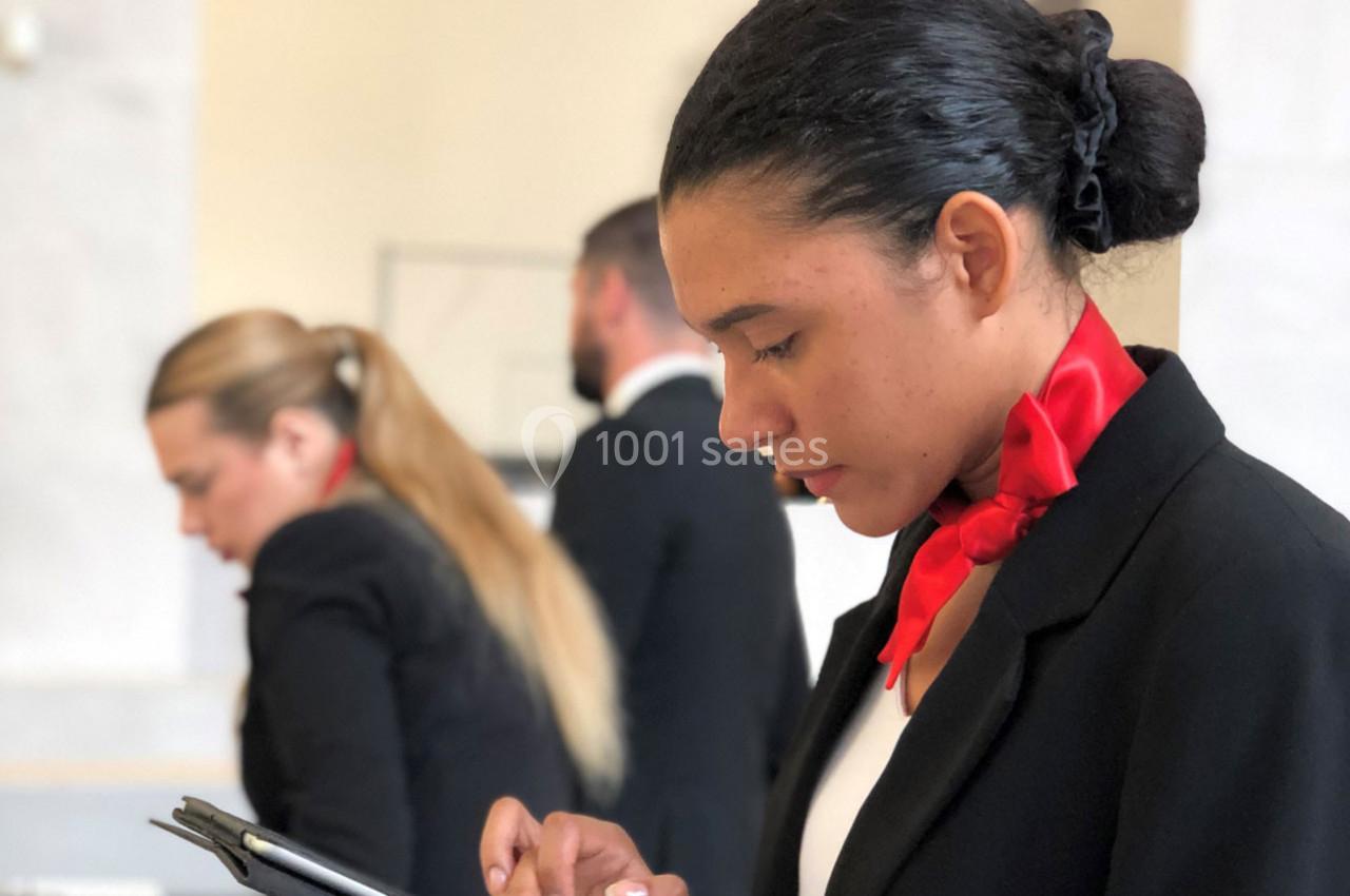 Une hôtesse en uniforme noir et foulard rouge consulte une tablette, avec deux collègues flous en arrière-plan.