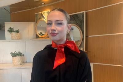 Femme souriante portant un béret rouge, debout près d'une vitrine décorée de fleurs colorées et de montres exposées.