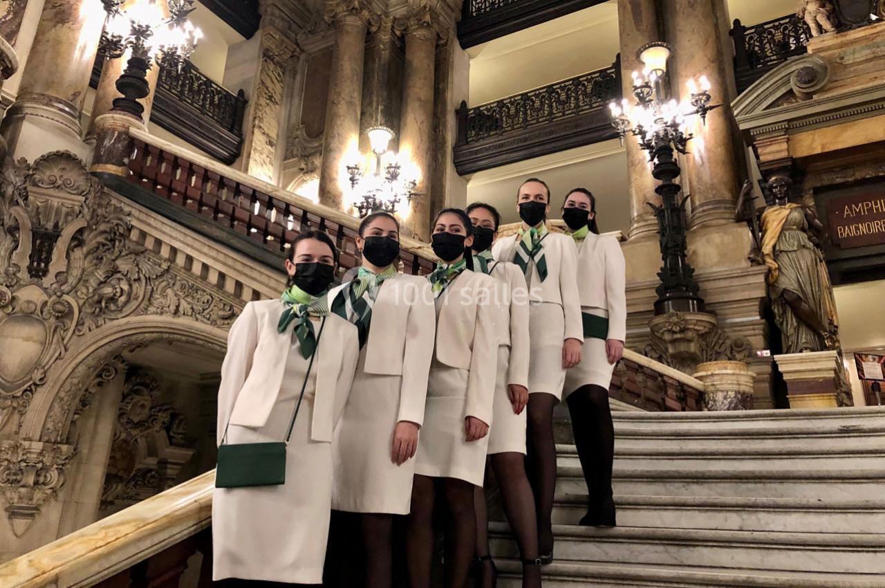 Cinq hôtesses en uniforme blanc posent sur un escalier décoré de sculptures et de lampes dans un bâtiment historique.