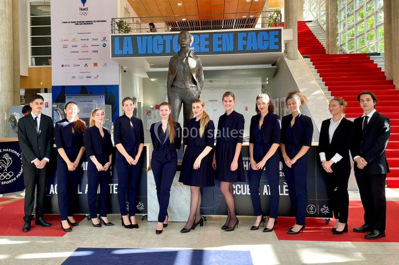 Un groupe de personnes en tenue formelle pose devant une statue et un escalier rouge dans un hall lumineux.