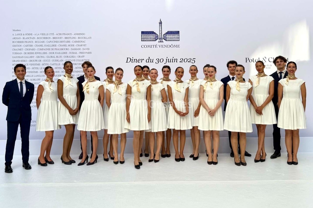 Photo MHBA - Mon Hôte-sse Bien Aimé-e #1 Un groupe de personnes posant en tenue élégante devant un mur blanc avec des inscriptions et un logo.