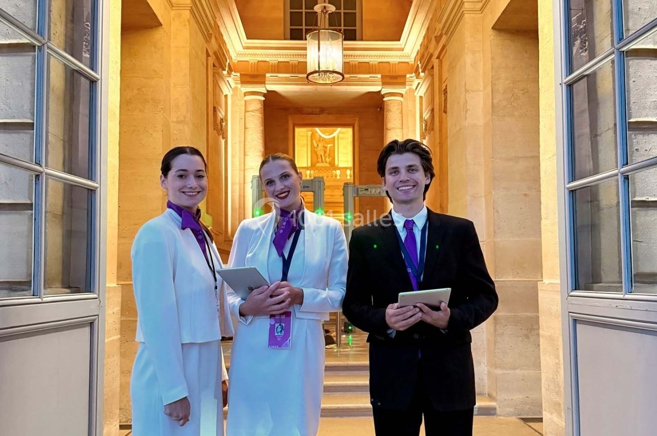 Photo MHBA - Mon Hôte-sse Bien Aimé-e #6 Trois hôtes souriants en tenue professionnelle, tenant des tablettes, devant une entrée lumineuse et élégante.