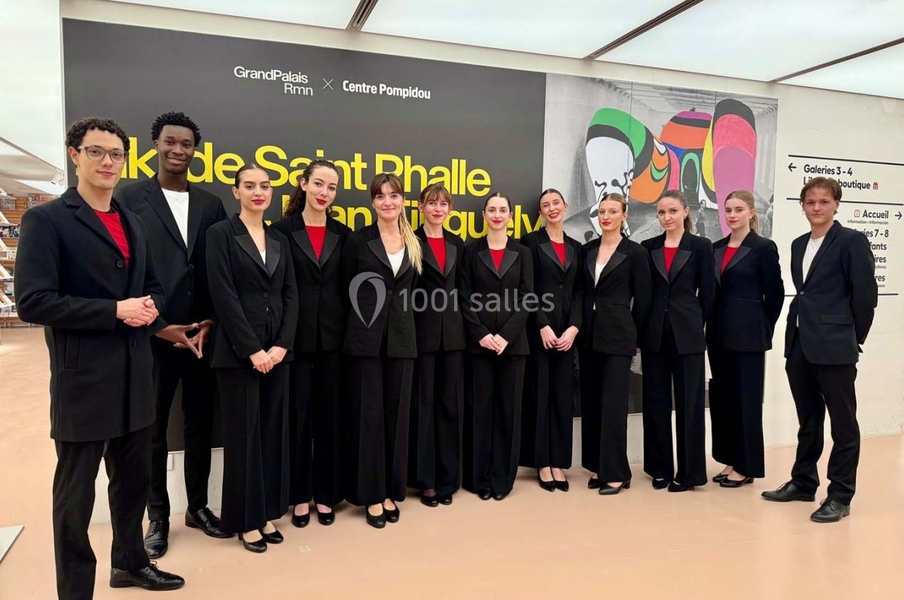 Photo MHBA - Mon Hôte-sse Bien Aimé-e #5 Un groupe de personnes en tenue formelle pose devant un mur affichant une exposition au Grand Palais Immersif.