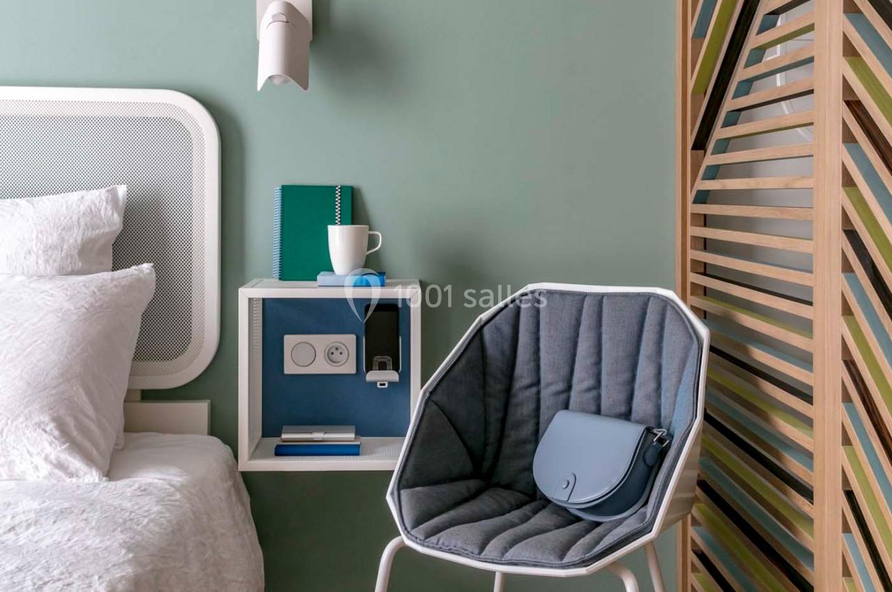 Coin de chambre avec lit, table de chevet intégrée, chaise grise avec sac bleu et cloison en bois colorée.