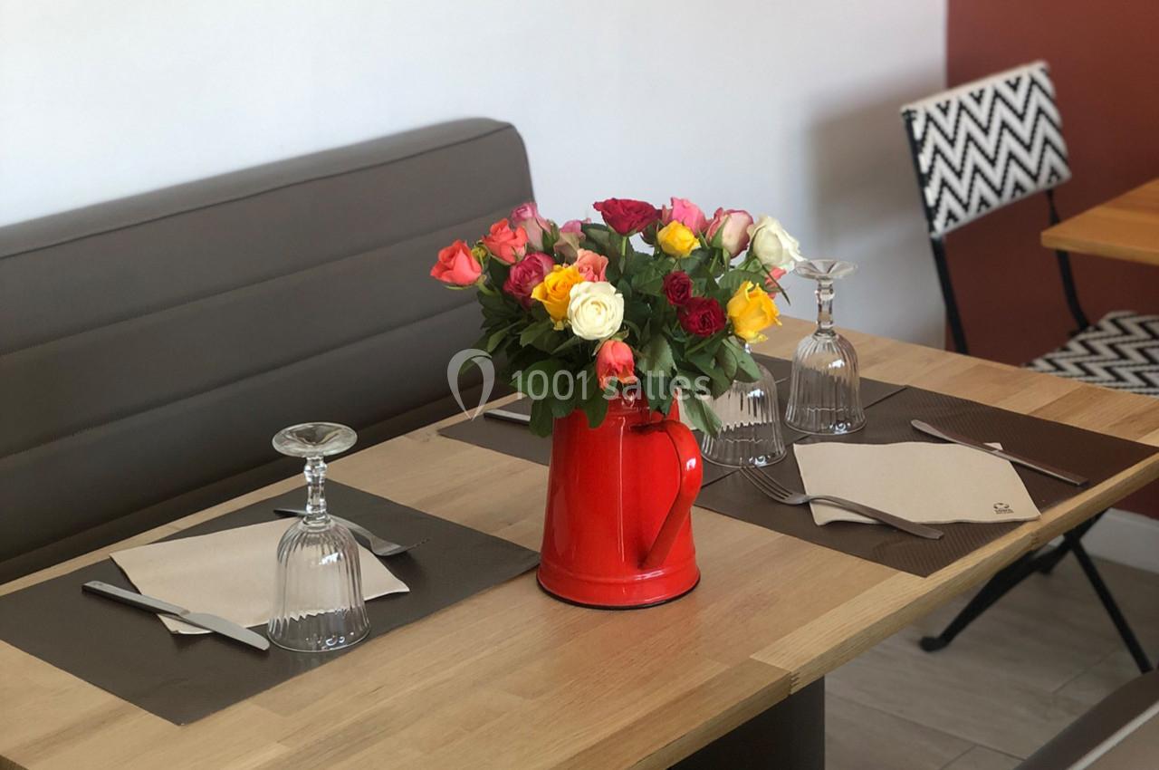 Table dressée avec un pichet rouge contenant des roses multicolores, entourée de verres et serviettes.