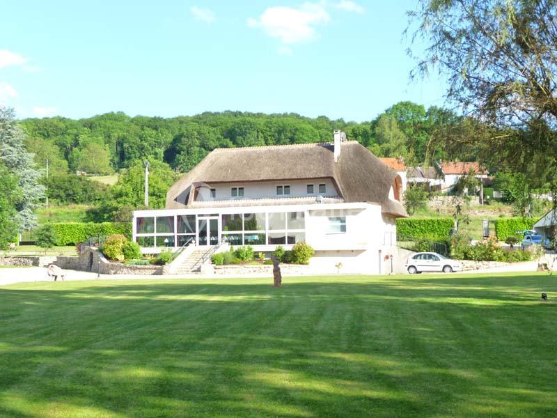 Maison avec toit de chaume entourée de verdure, située dans un paysage rural avec pelouse et arbres.
