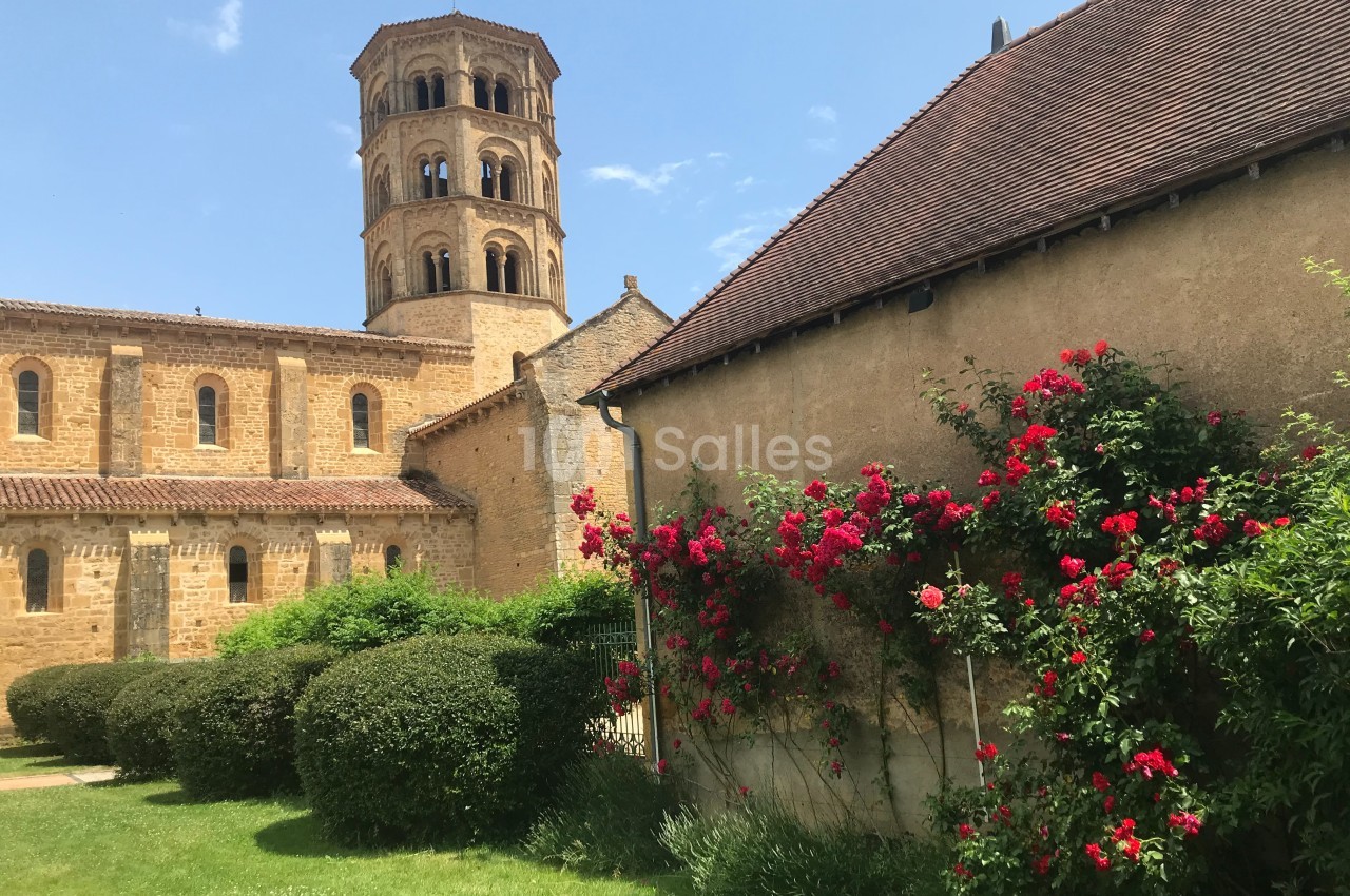 Église en pierre avec clocher roman, jardin fleuri et rosiers rouges grimpant sur un mur sous un ciel bleu.