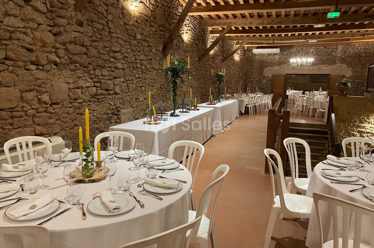 Salle en pierre aménagée pour un repas, avec tables rondes dressées et tables rectangulaires en fond.