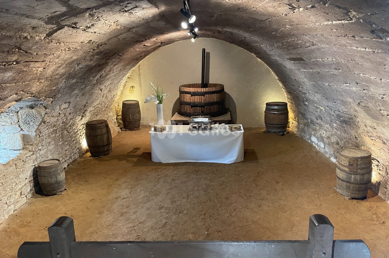 Salle voûtée en pierre avec un pressoir ancien, des tonneaux en bois et une table dressée au centre.