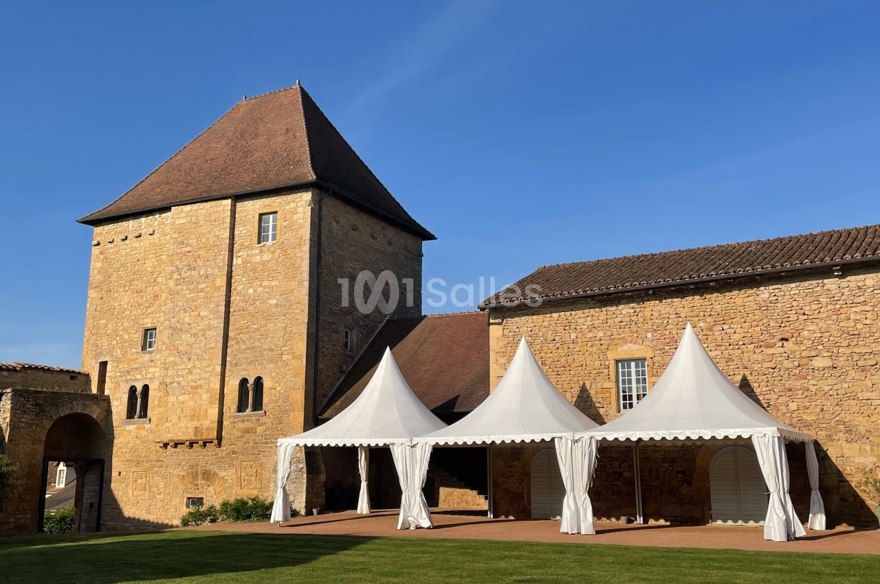 Tour en pierre historique avec toit en pente, entourée de tentes blanches installées sur une pelouse sous un ciel bleu.