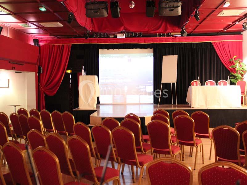Salle de conférence avec des chaises rouges alignées, une scène équipée d'un écran, d'un tableau et de tables blanches.