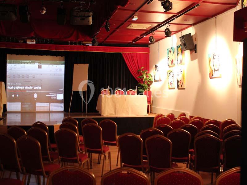 Salle de conférence vide avec des rangées de chaises rouges, une table blanche, un écran de projection et des tableaux…
