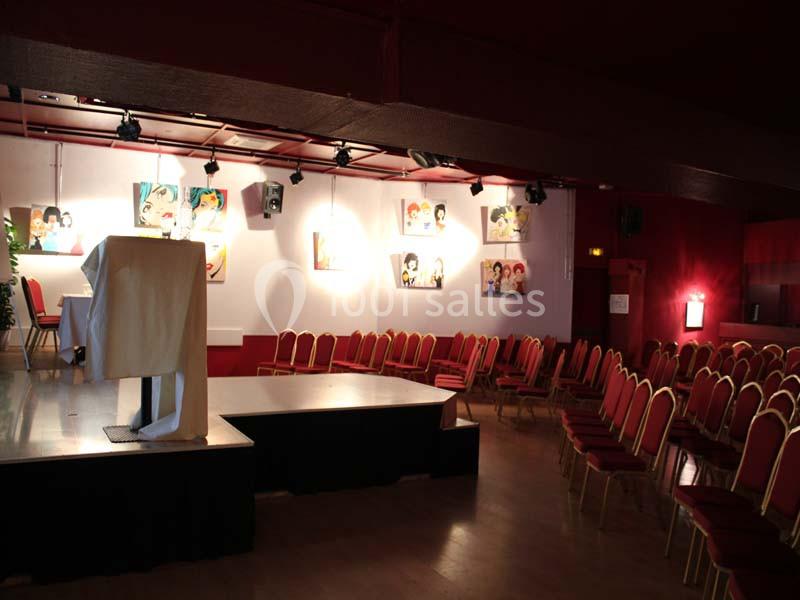 Salle de conférence avec des rangées de chaises rouges, une estrade et des tableaux colorés accrochés au mur.