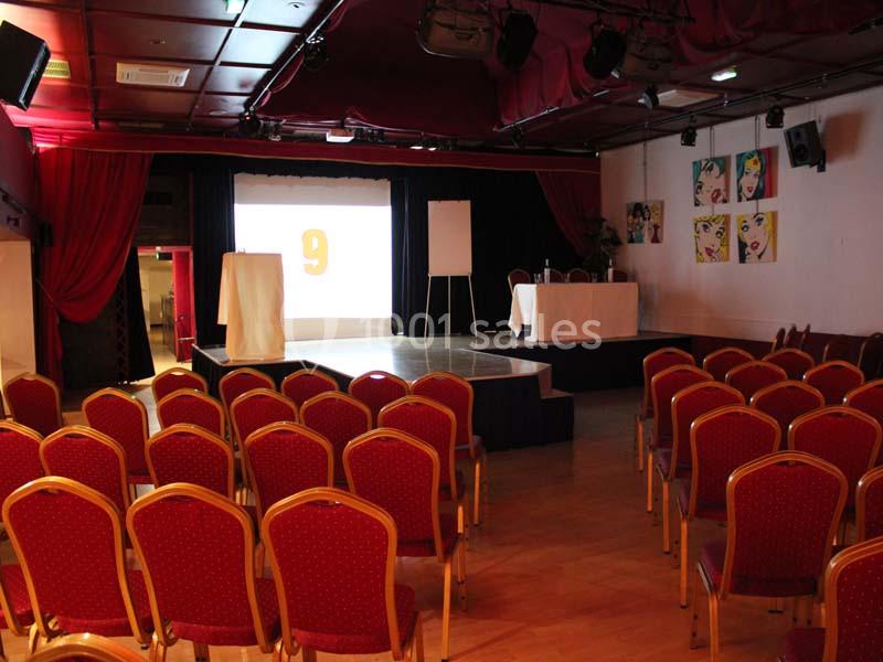 Salle de conférence avec des rangées de chaises rouges, une scène équipée d'un écran et d'un pupitre.