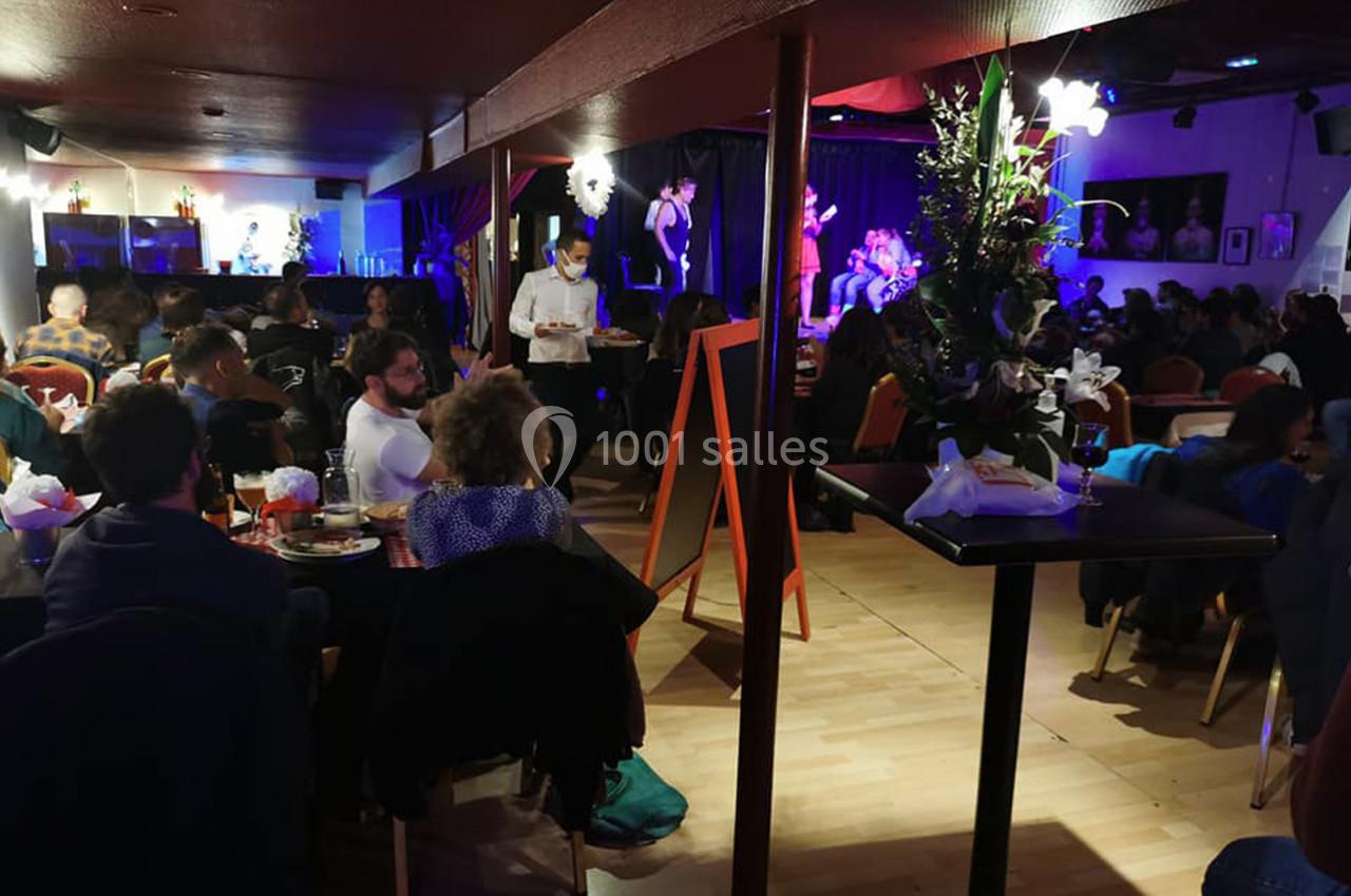 Salle animée avec des tables occupées, un serveur en mouvement et une scène où des musiciens jouent sous des lumières…