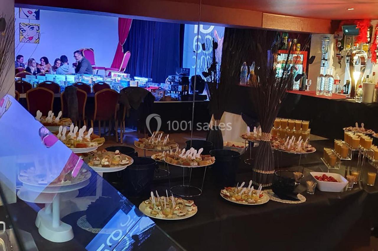 Buffet de verrines et amuse-bouches disposé dans une salle avec tables, chaises et décorations lumineuses en arrière-plan.