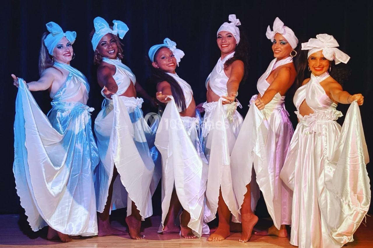 Six femmes en tenue blanche traditionnelle dansent sur scène, tenant des pans de tissu, devant un fond sombre.