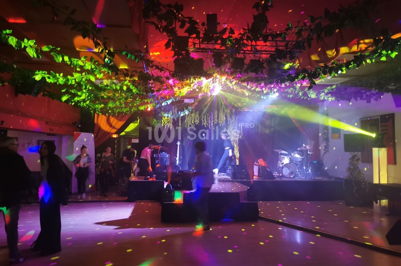 Salle de fête décorée de guirlandes lumineuses et végétales, avec piste de danse éclairée et scène musicale en arrière-plan.